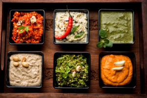 Six different chili dip sauces in black square serving dishes on a wooden tray.