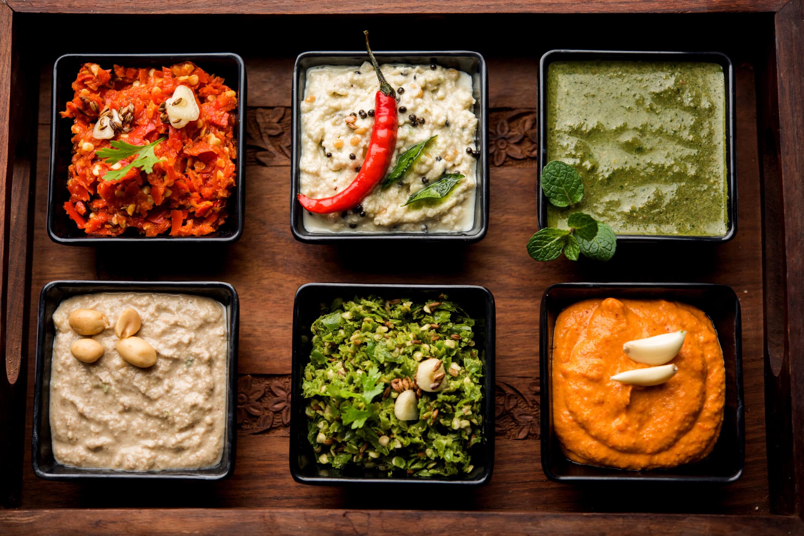 Kuusi erilaista chilistä dippikastiketta mustissa neliönmuotoisissa tarjoiluastioissa puisella alustalla.