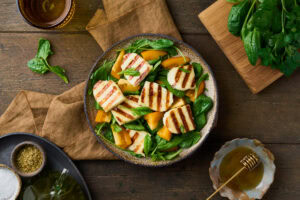 Top-down view of a halloumi salad with spinach and tomatoes. Honey and spices are placed on a wooden table alongside the dish.