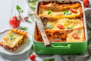 A cheesy lasagna in a baking dish, with a slice lifted onto a white plate; cherry tomatoes and basil leaves arranged around it.