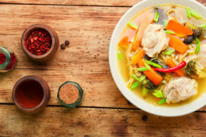 A bowl of steaming spicy chicken soup with vegetables and chili. On the wooden table, small bowls with various spices.