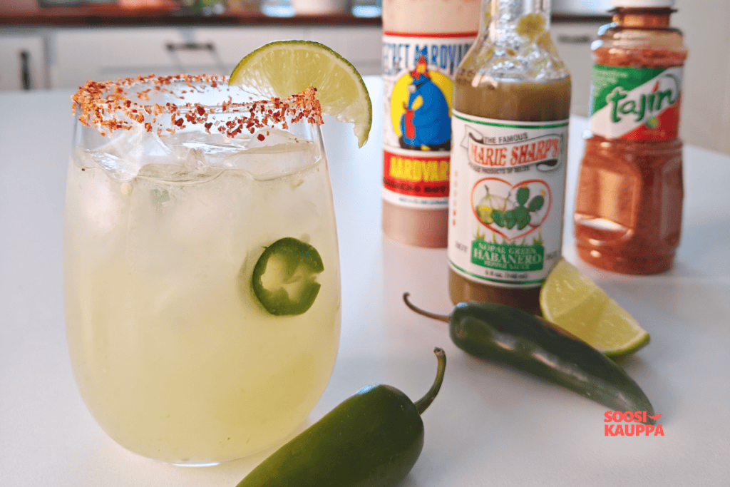 Valmis Spicy Margarita jäillä; lasin reunassa Tajín-maustetta ja limelohko, taustalla chilikastikkeita ja jalapeñoja.