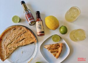 Vegaaninen parsapiirakka pöydällä ylhäältä päin kuvattuna ja piirakkapalanen lautasella. Pöydällä lisäksi chilikastikkeita, sitruuna, limejä ja kylmiä juomia.