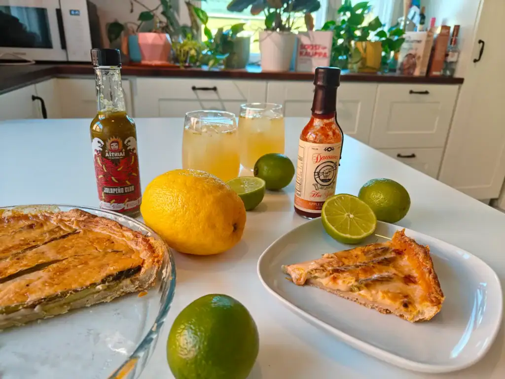A close-up of an Easter serving suggestion, with asparagus pie, hot sauces, and cold drinks surrounded by citrus fruits on the table. In the background, light kitchen cabinets, houseplants, and a Soosikauppa package. 