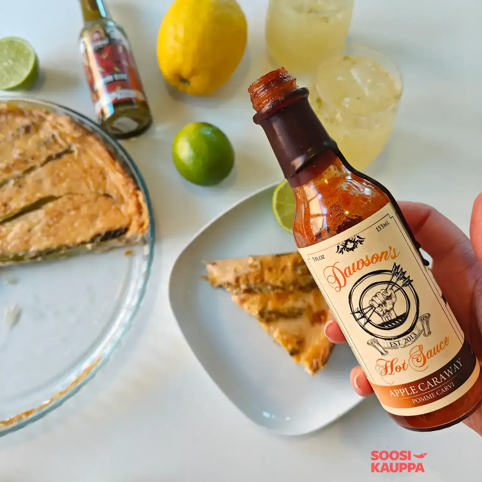 Dawson's Apple Caraway Hot Sauce bottle in hand, with a vegan asparagus pie in the background, a slice of pie on a plate, lemons, limes, and cold drinks.