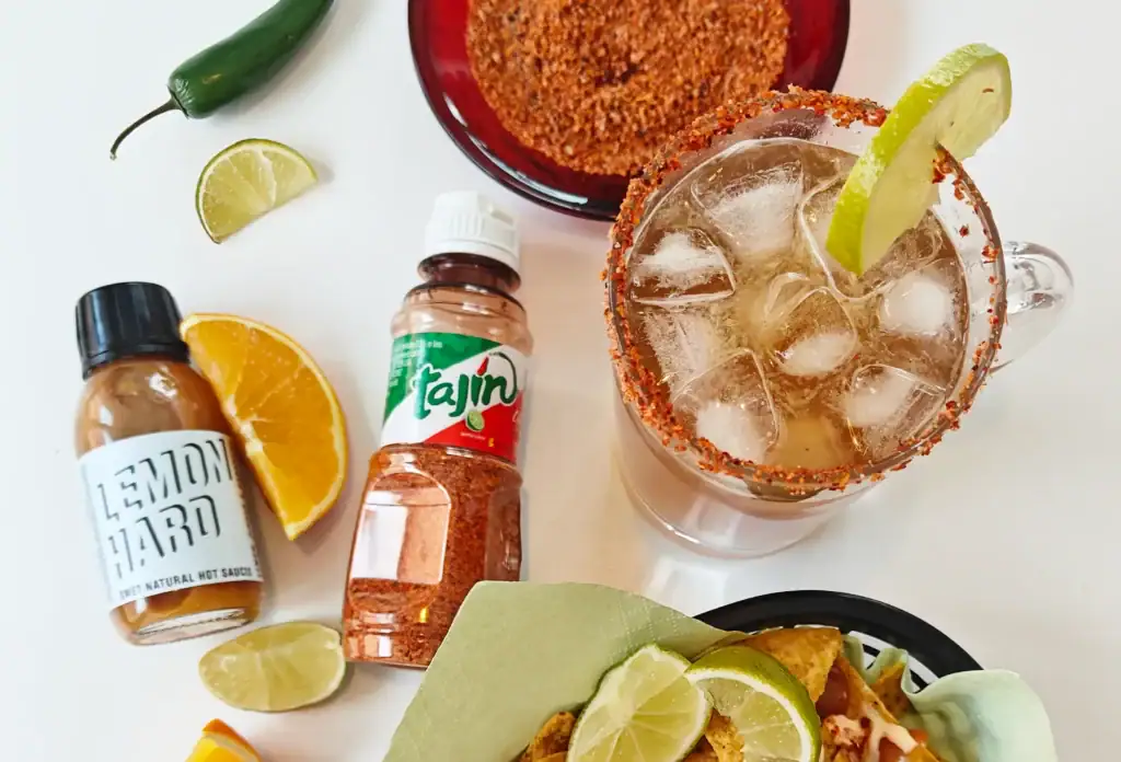 Michelada-olutdrinkki lasissa, jonka reunalla Tajín-mauste ja limelohko, ympärillä chilikastikkeita ja sitruksia.