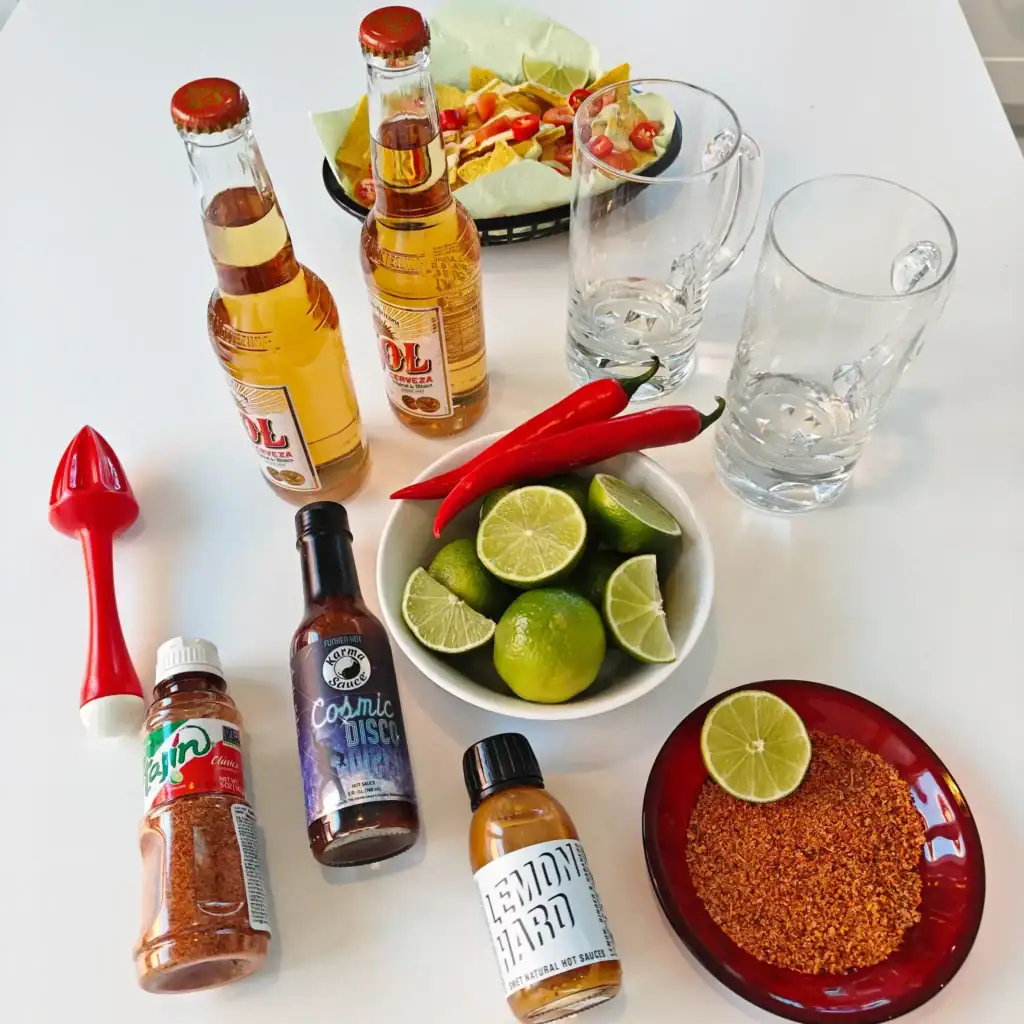 Micheladaan tarvittavat ainekset pöydällä: lager, lime, chilikastikkeet ja Tajín-mauste.