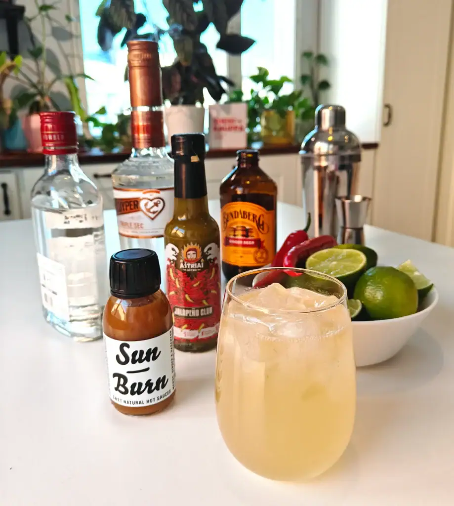 Moscow Mule -drinkki lasissa, taustalla vodka, limejä, chilejä, inkivääriolut ja Soosikaupan chilikastikkeet.