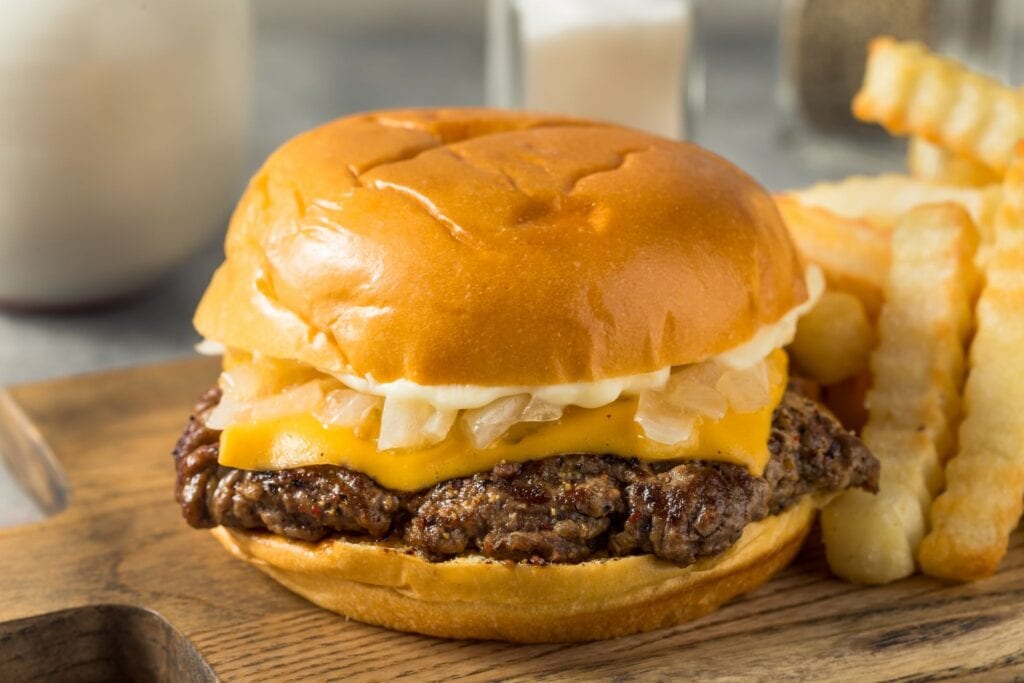 Yksinkertainen smashburger, jossa välissä pihvi, juustoa ja sipulia, vieressä ranskalaisia tarjoiltuna puisen leikkuulaudan päältä. 